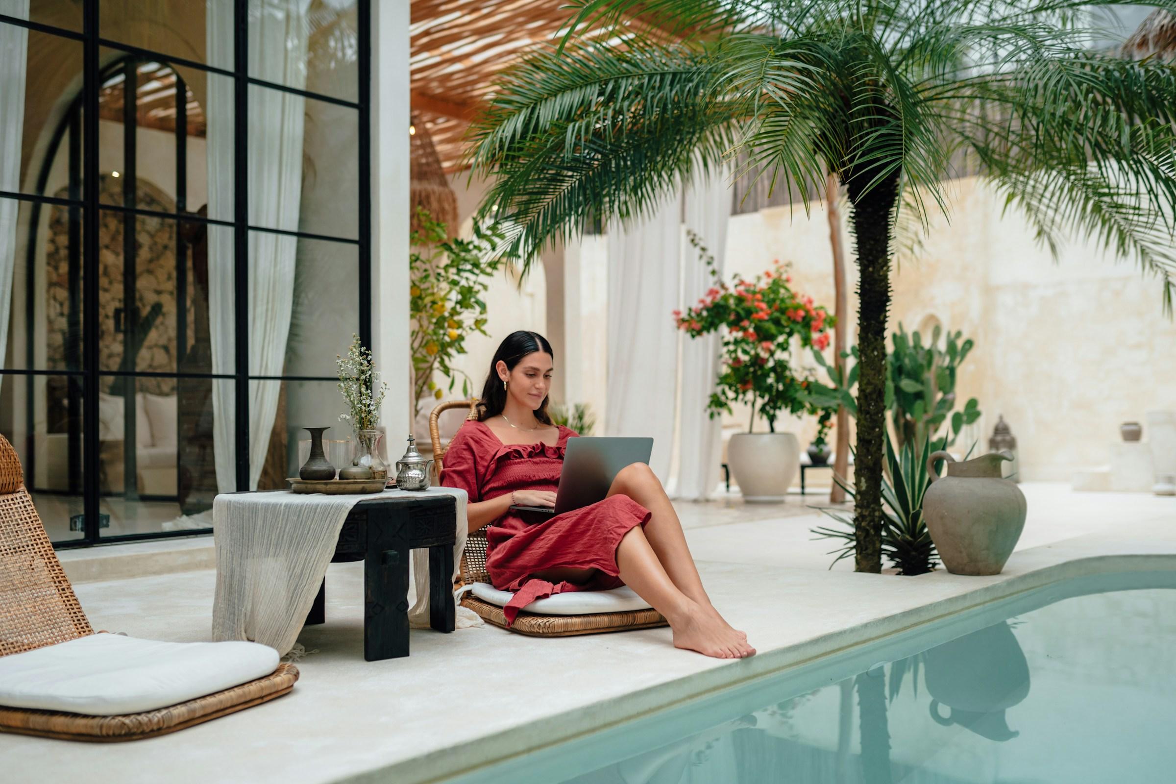 Woman using a laptop by a pool with palm trees and indoor plants in the background