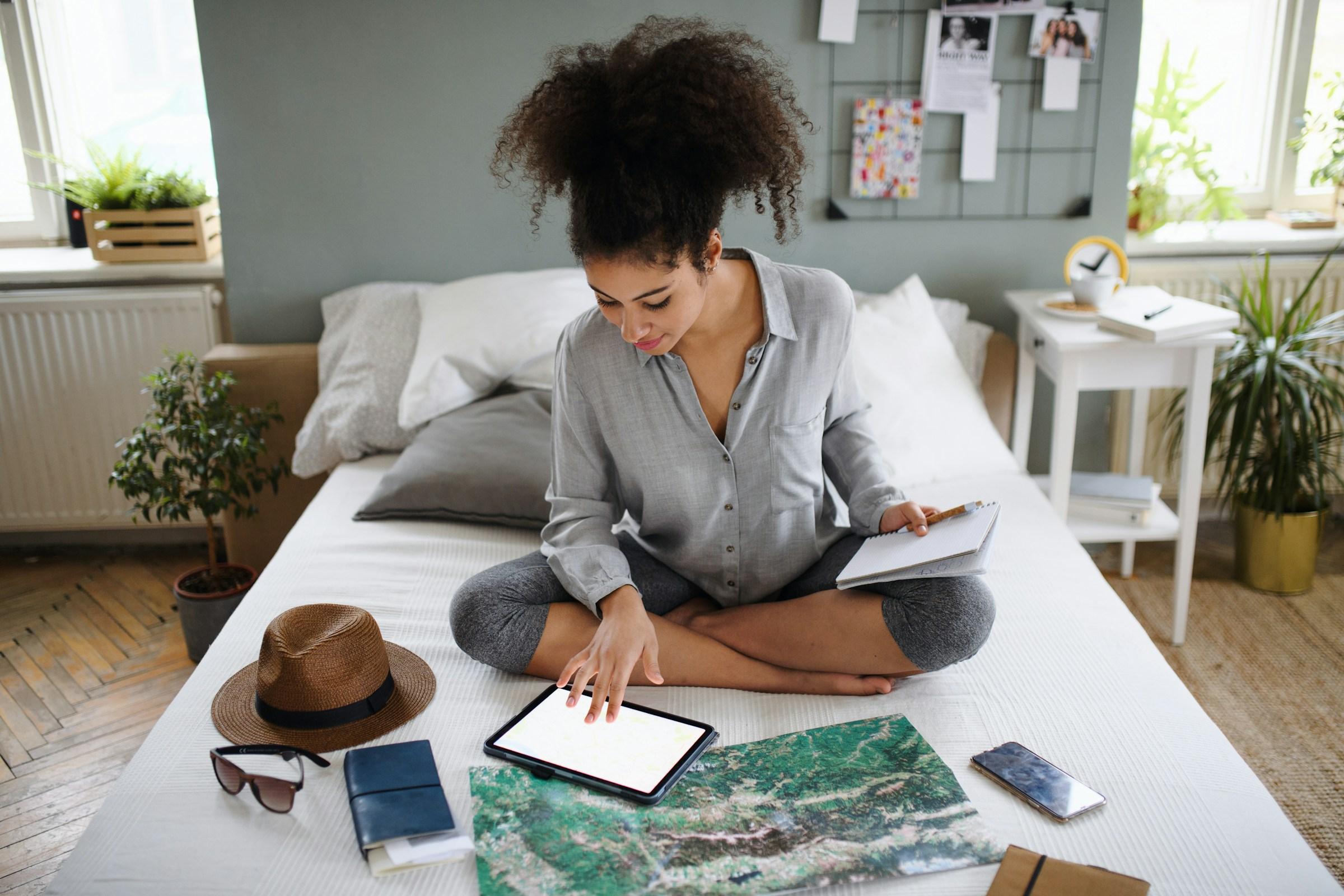 Woman sitting on a bed with travel items and a tablet, in a home setting.